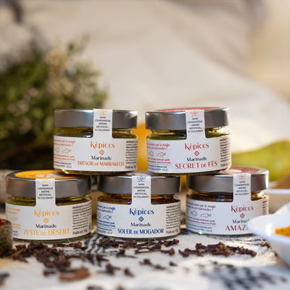 Mise en scène du Coffret Découverte Képices – cinq marinades marocaines artisanales empilées parmi des épices et herbes aromatiques.