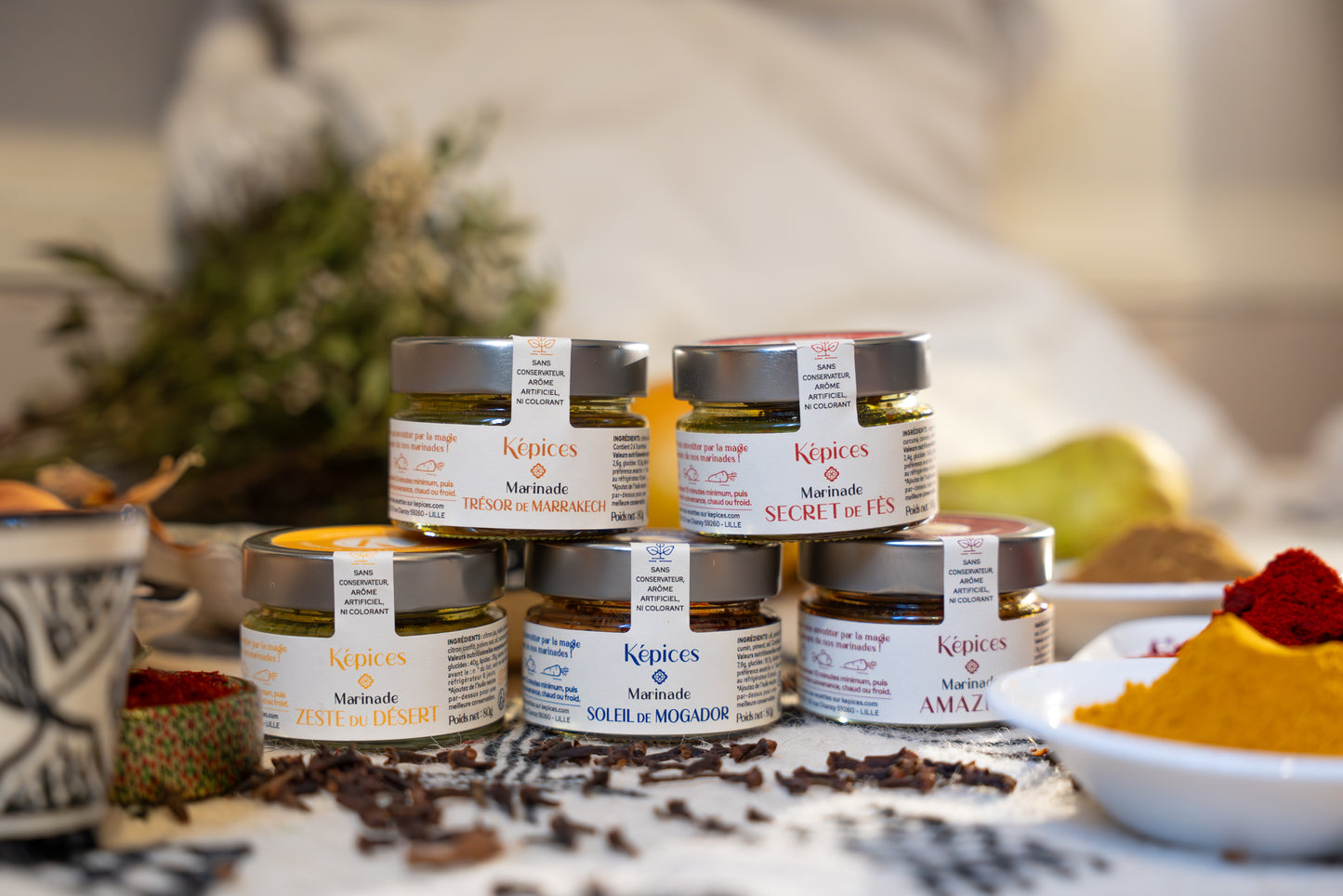 Mise en scène du Coffret Découverte Képices – cinq marinades marocaines artisanales empilées parmi des épices et herbes aromatiques.