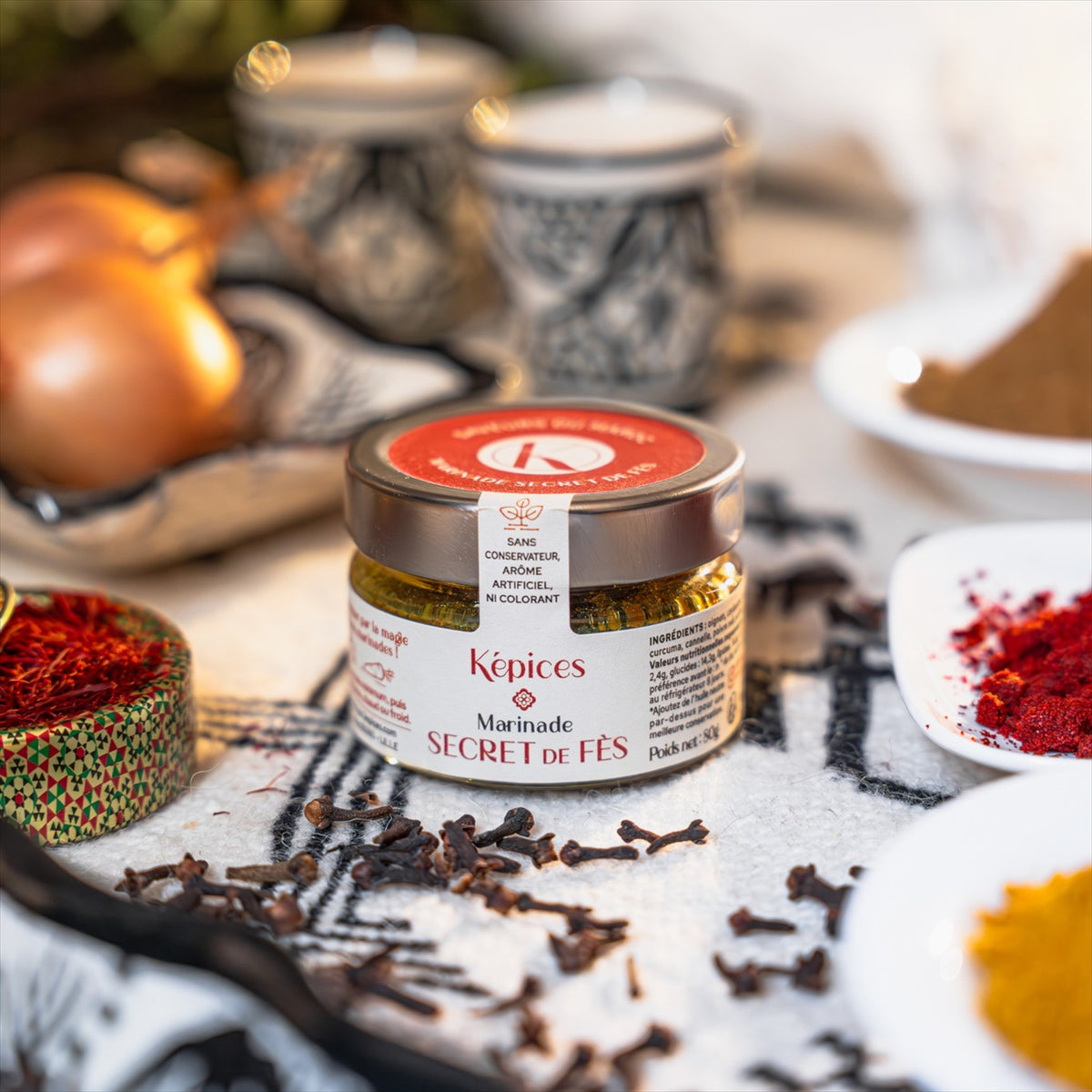 Marinade Secret de Fès Képices posée sur un tissu oriental avec poudre de curcuma et ustensiles en métal.