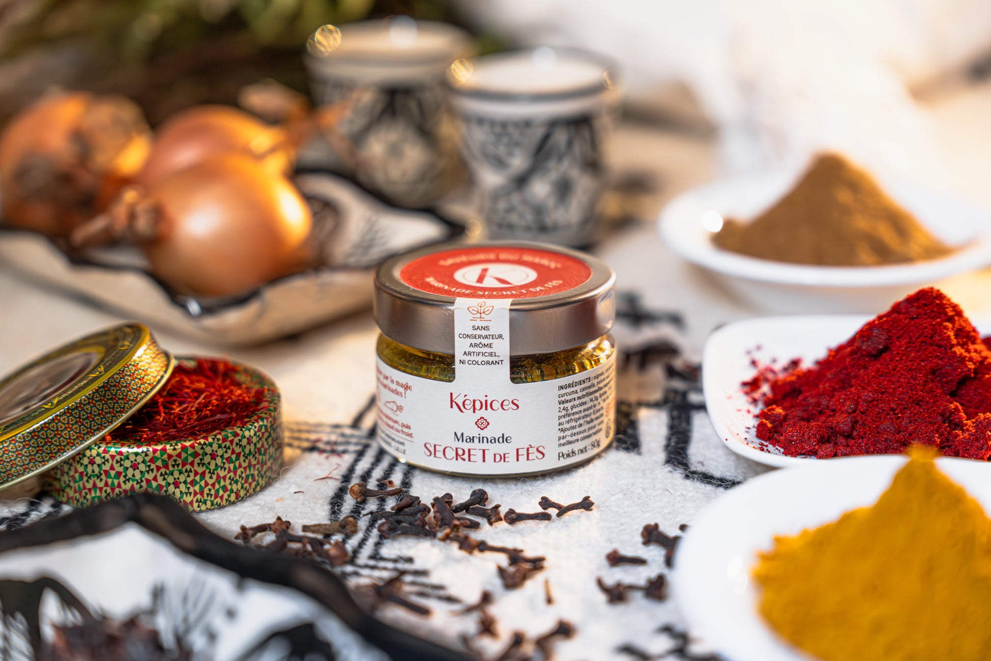 Marinade Secret de Fès Képices posée sur un tissu oriental avec poudre de curcuma et ustensiles en métal.